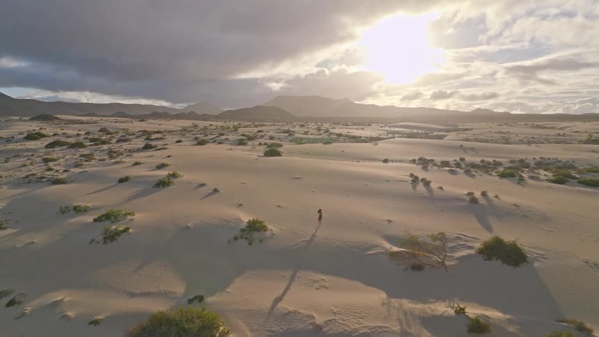 An epic aerial view follows a lone woman walking through a vast desert landscape of sand dunes at sunset
