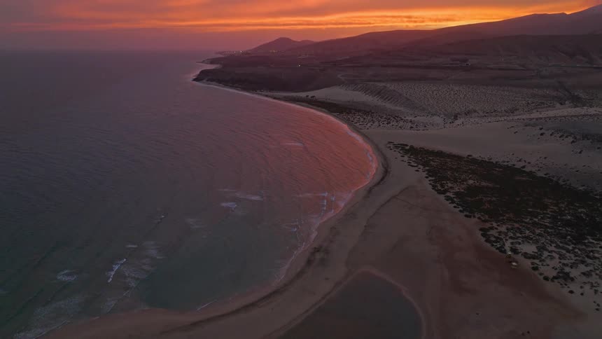 An aerial view tilts up to reveal a fiery orange sunset reflecting on the ocean along a vast desert coastline