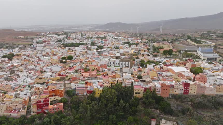 An aerial view slowly pans across a colorful Spanish town, revealing its historic church and the surrounding arid landscape with mountains and wind turbines