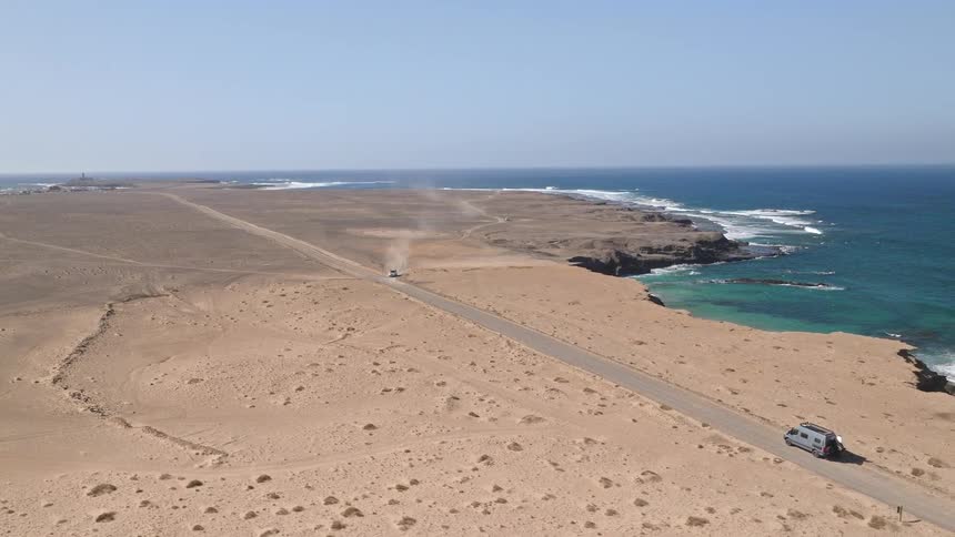 An aerial view shows a lone camper van driving down a scenic desert road that runs along a beautiful turquoise coastline