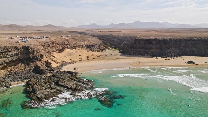 Aerial view flying away from a beautiful secluded beach cove with turquoise water, surfers, and a vast desert landscape
