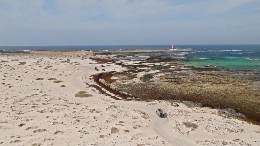 An aerial shot follows a camper van driving on a sandy road through a coastal desert towards a beautiful lighthouse