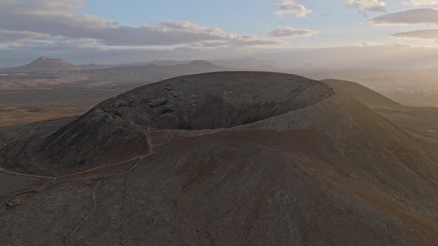 A stunning aerial view orbits a massive, dormant volcano crater as the sun rises over a vast and barren landscape