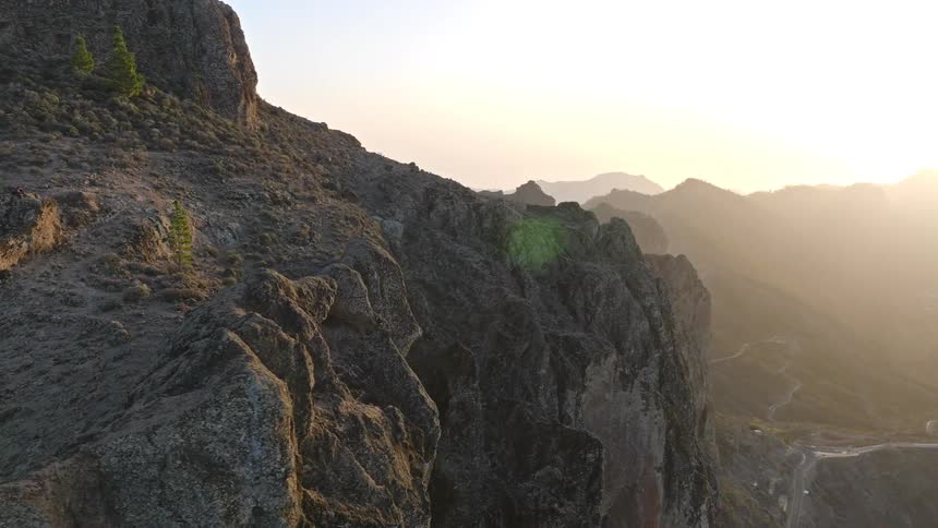 A cinematic drone shot pulls back from a unique natural rock arch, revealing a vast and hazy mountain landscape at golden hour