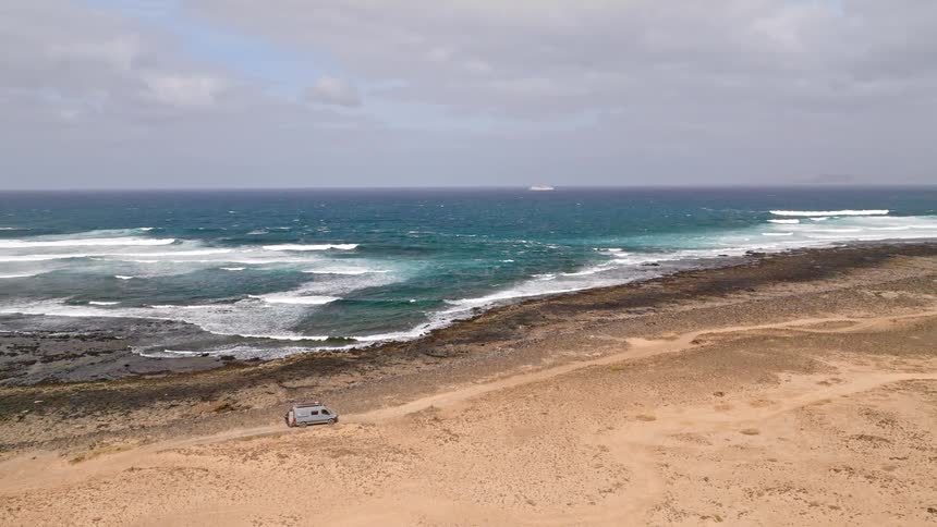 An aerial view shows a camper van driving on a dirt road along the wild, rocky coast of a volcanic island, with turquoise waves breaking on the shore