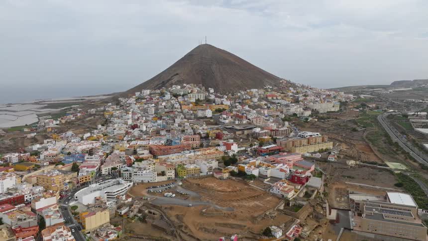 An expansive aerial shot slowly pulls back to reveal the town of Gáldar clinging to the slopes of a prominent mountain on Gran Canaria island