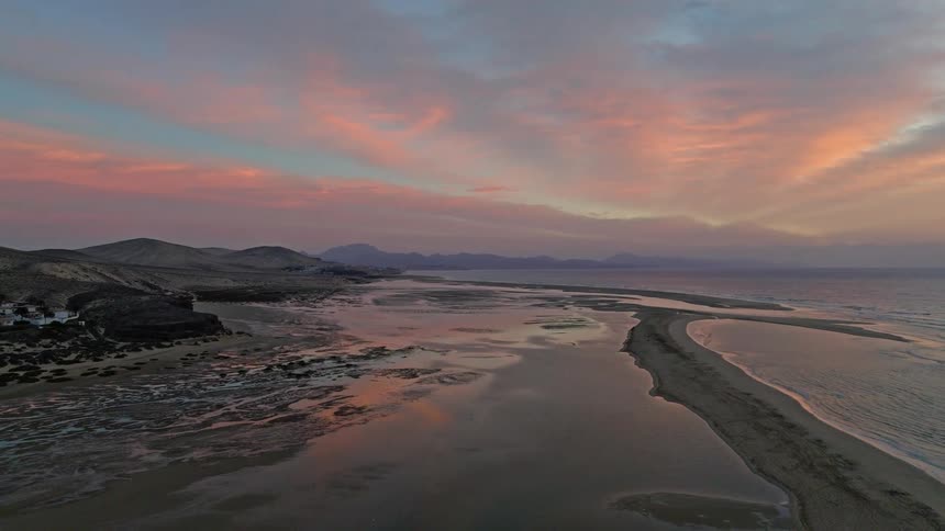 Stunning aerial view pulling back over a calm tidal lagoon reflecting a vibrant pink and orange sunset along a rugged, mountainous coastline