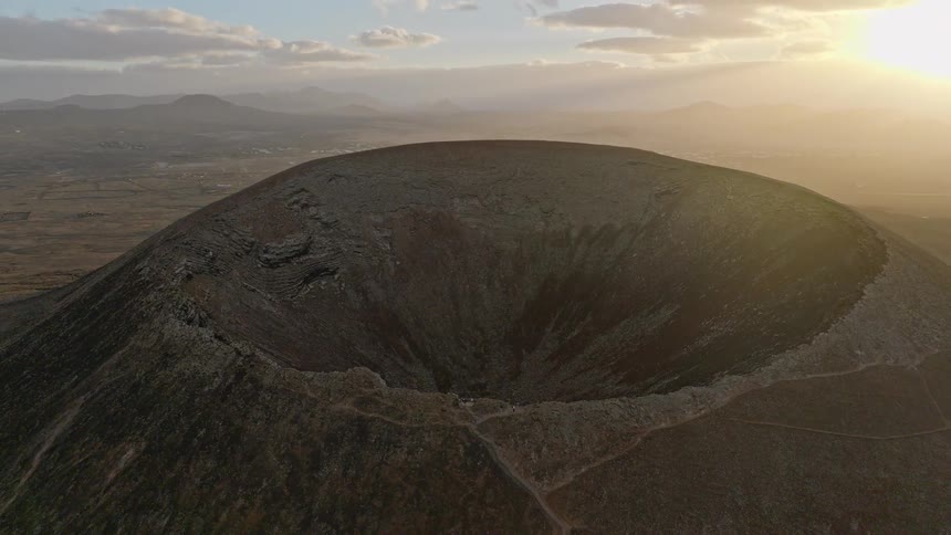 An epic aerial shot reveals the massive crater of a dormant volcano as the camera pulls back to showcase the vast landscape at golden hour