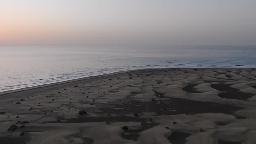 A beautiful aerial time-lapse captures the sun rising over the calm ocean, illuminating the vast, shadowy sand dunes on the coast