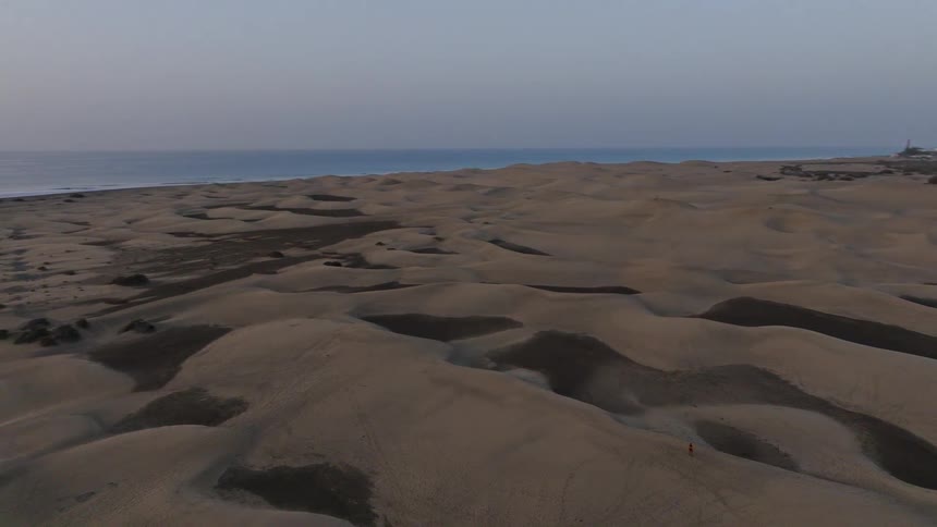 An epic aerial pullback shot reveals a woman walking through a vast desert of sand dunes along the coastline at sunrise