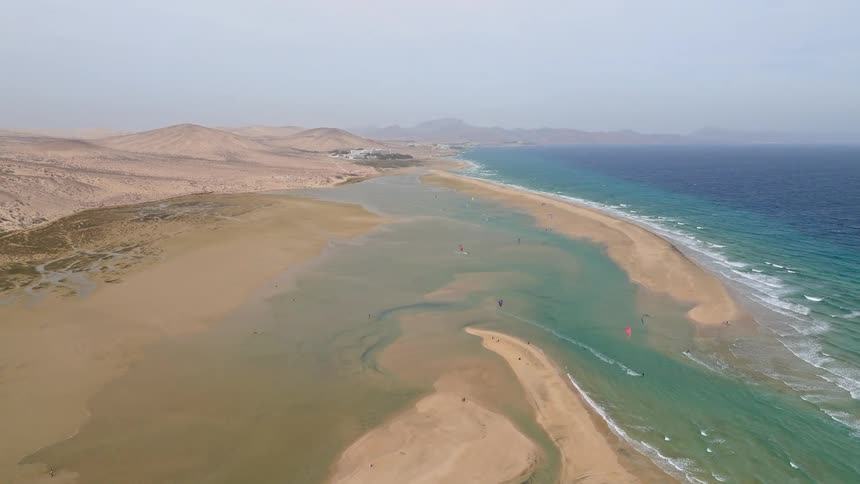 A slow aerial pullback reveals a stunning desert coastline where kitesurfers glide across a shallow turquoise lagoon