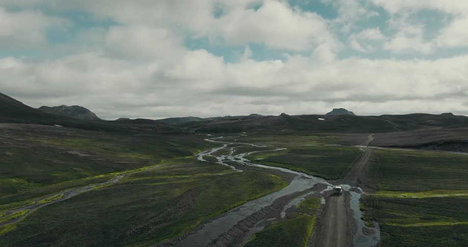 An aerial shot follows a 4x4 vehicle as it drives on a remote dirt road and crosses a river in a vast, green valley