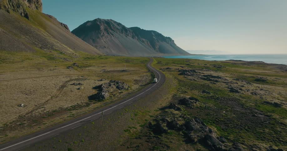 Aerial shot of a white car driving on a winding coastal road through a vast, mountainous landscape in Iceland
