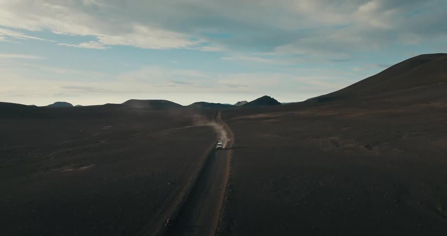 An epic aerial shot follows a lone white 4x4 vehicle driving on a dusty dirt road through a vast, dark, and desolate volcanic landscape