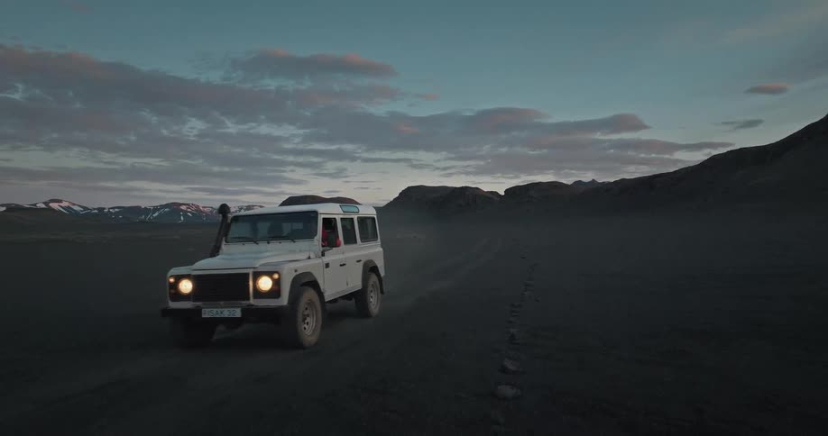 A rugged white SUV slowly drives across a vast and desolate volcanic black sand desert at dusk in the Icelandic highlands