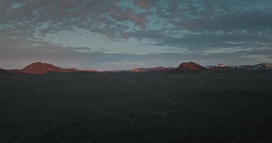Aerial footage glides over a vast and desolate rocky landscape toward distant mountains basking in the warm glow of a dramatic sunset