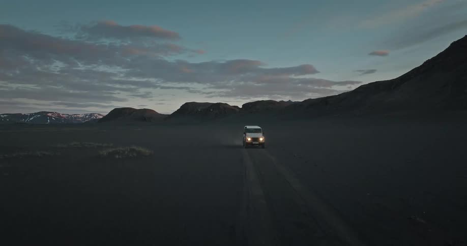 An aerial shot follows a white van driving through a dark, remote volcanic landscape in Iceland at dusk