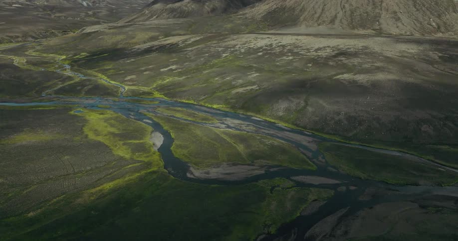 A wide aerial shot shows clouds moving over a green valley with a braided river running through it in the highlands of Iceland