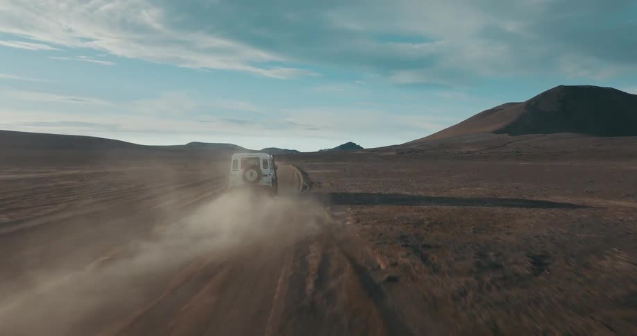 An aerial drone shot follows a white off-road vehicle as it kicks up dust on a rugged road through a vast, desolate landscape