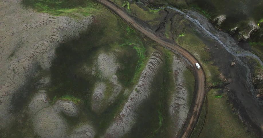 A top-down aerial view follows a white camper van as it navigates a winding dirt road through a vast, moss-covered volcanic landscape