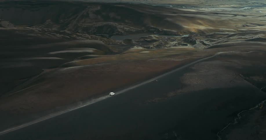 Epic aerial shot of a white SUV driving on a remote dirt road through the desolate volcanic highlands of Iceland