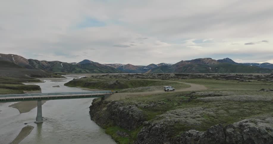 An aerial view shows a white 4x4 vehicle crossing a bridge over a river and driving on a dirt road in the epic Icelandic Highlands
