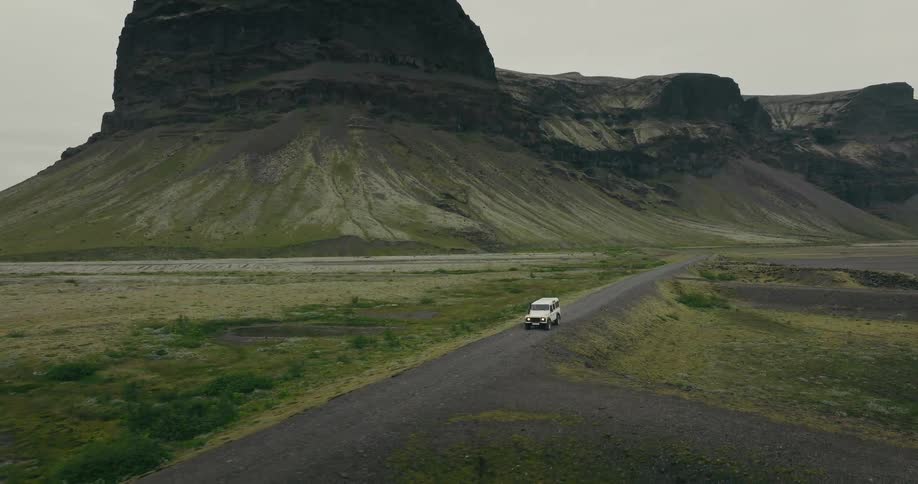 Aerial footage shows a white 4x4 vehicle driving on a remote gravel road through the vast, mountainous highlands of Iceland