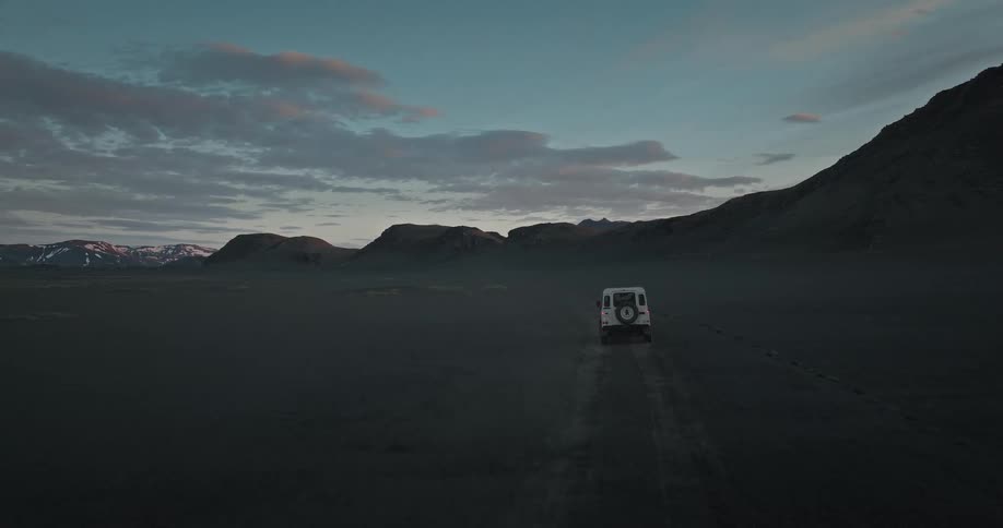 A white off-road vehicle drives through a vast, desolate black sand desert landscape at twilight, as seen from an aerial perspective