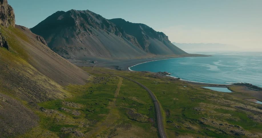 Cinematic aerial shot of a car traveling on a scenic coastal highway nestled between dramatic mountains and the serene blue ocean in Iceland