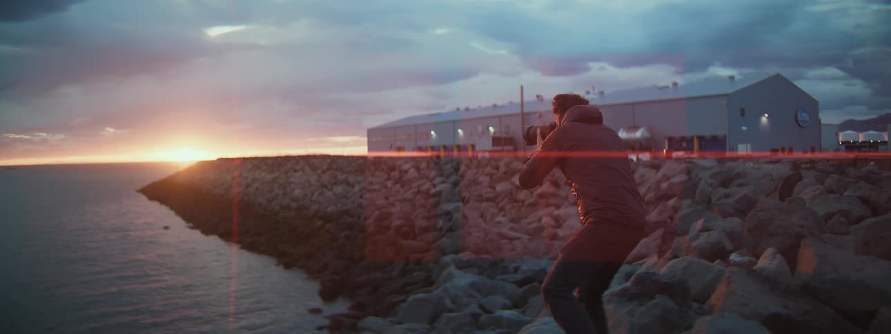 A photographer stands on a rocky jetty taking photos of the ocean during a beautiful sunset with a cinematic lens flare