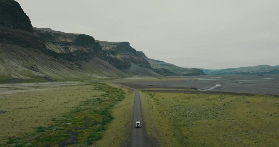 A high-angle aerial shot follows a white SUV driving on a long, desolate road through the epic, green landscapes of a mountainous valley in Iceland