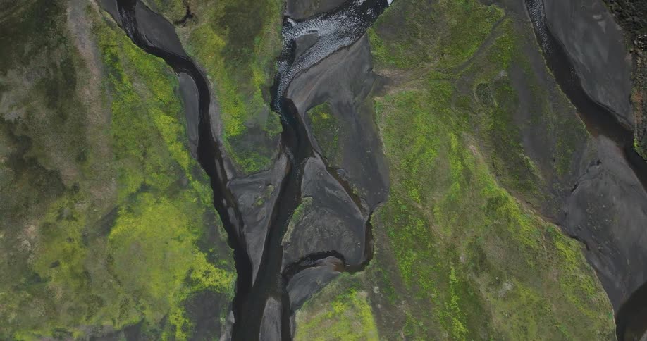A stunning top-down aerial shot slowly rises above an intricate braided river system flowing through a vibrant green, moss-covered volcanic valley