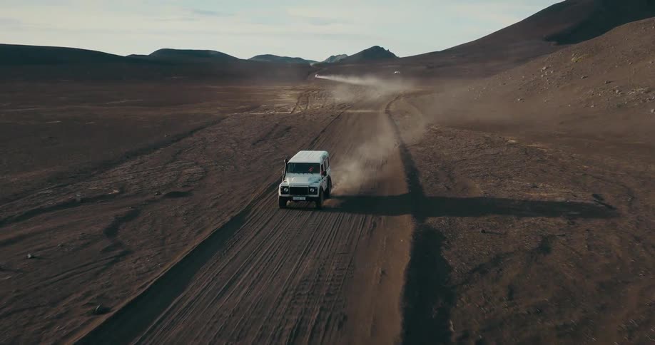 An aerial view follows a white SUV kicking up dust as it drives along a remote road in a vast, barren desert landscape