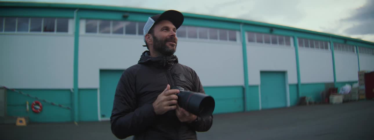 A male photographer wearing a hat and jacket holds a camera while talking and gesturing outdoors in slow motion