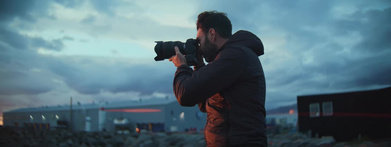 A male photographer in a dark jacket takes pictures at twilight in a rocky, industrial landscape
