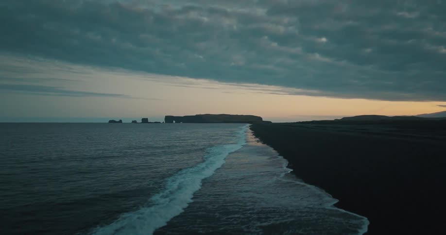 A moody aerial shot flies backward over gentle waves washing onto a black sand beach at twilight