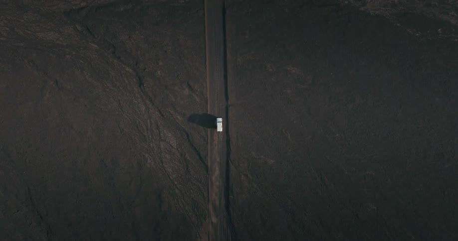 An aerial top-down view follows a lone white van driving on a straight road through a vast, dark, and desolate landscape