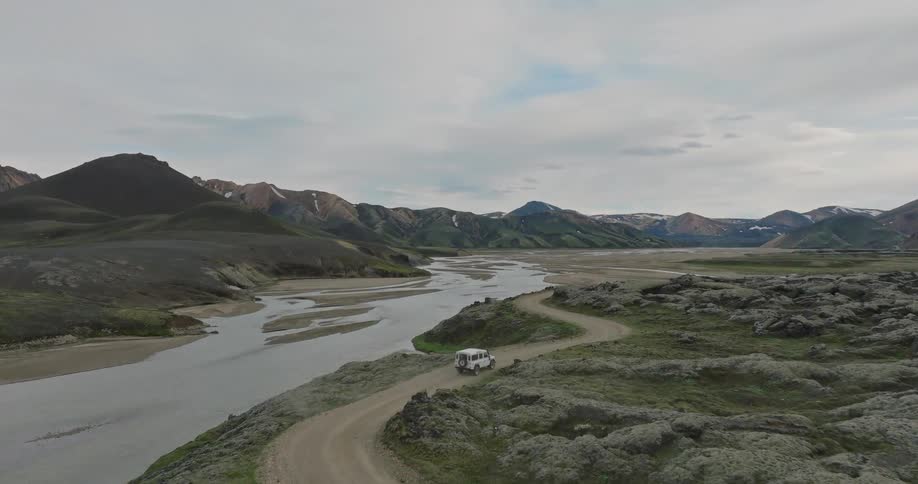 An aerial view follows a white 4x4 vehicle on a remote dirt road alongside a river in the vast, mountainous highlands of Iceland