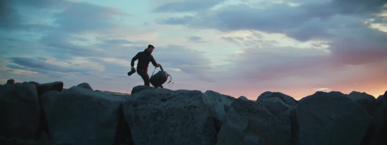 A male photographer carefully walks across a rocky jetty with his camera and backpack to set up for a sunset shot