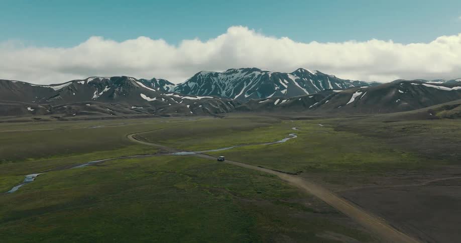 Epic aerial footage tracks a lone 4x4 vehicle driving through a vast, wild green valley towards majestic snow-capped mountains in Iceland
