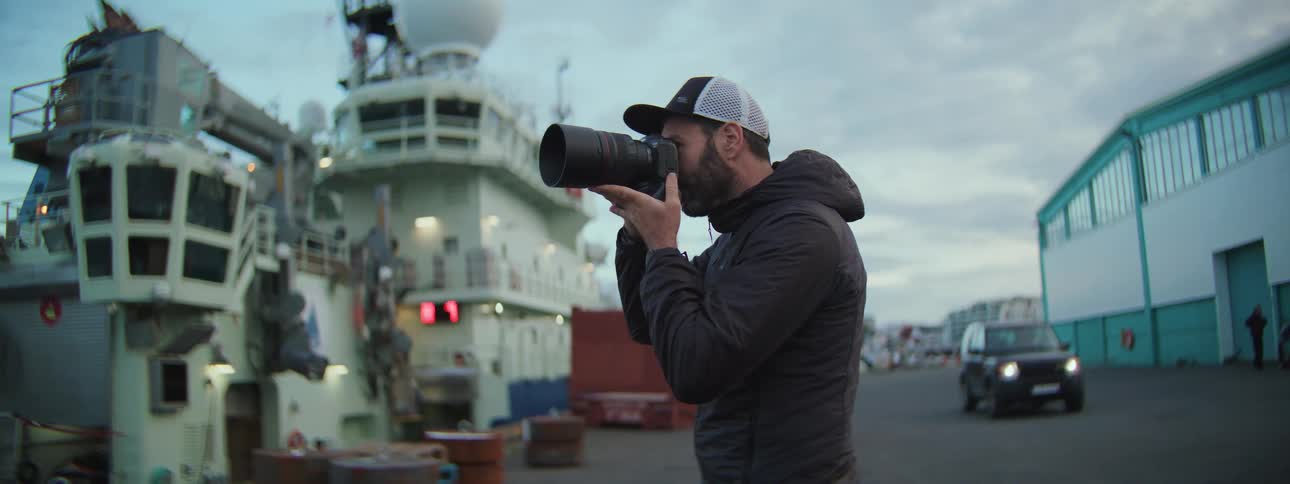 A photographer with a telephoto lens captures images of a large ship at an industrial harbor at dusk