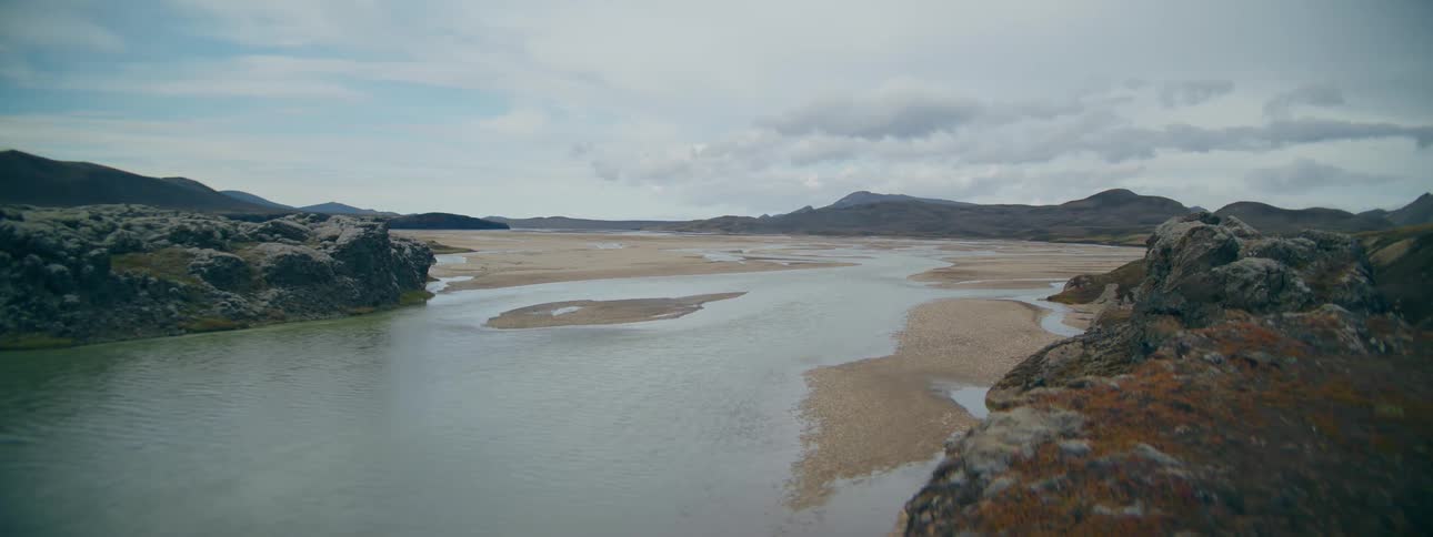 A tranquil river flows through a vast, remote, and rocky Icelandic landscape under a cloudy sky