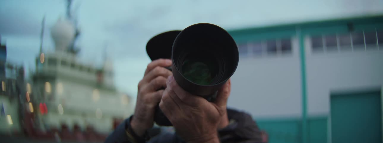 A photographer with a large telephoto lens aims, then lowers his camera to survey the scene at a harbor