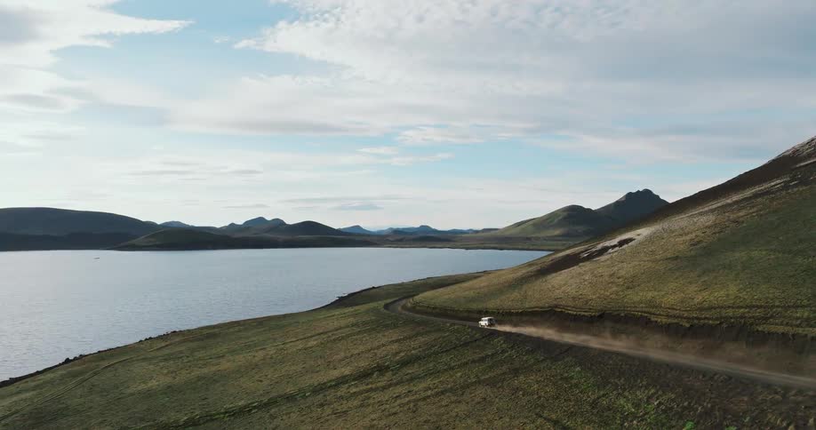 A white 4x4 vehicle kicks up dust as it drives along a remote dirt road next to a vast lake and rolling green hills