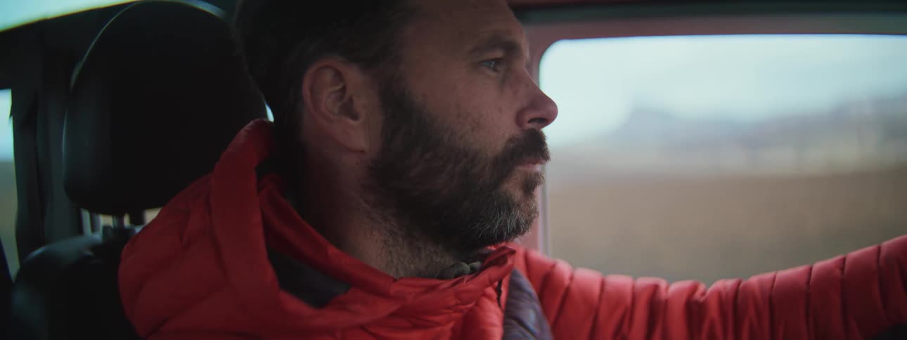 Close-up profile of a bearded man in a red puffer jacket intently driving a vehicle through a rural landscape