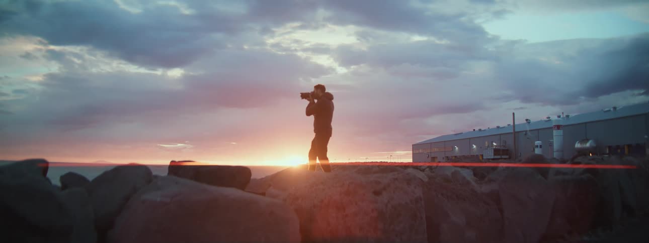 A silhouetted photographer stands on a rocky jetty, capturing the beautiful sunset over the ocean
