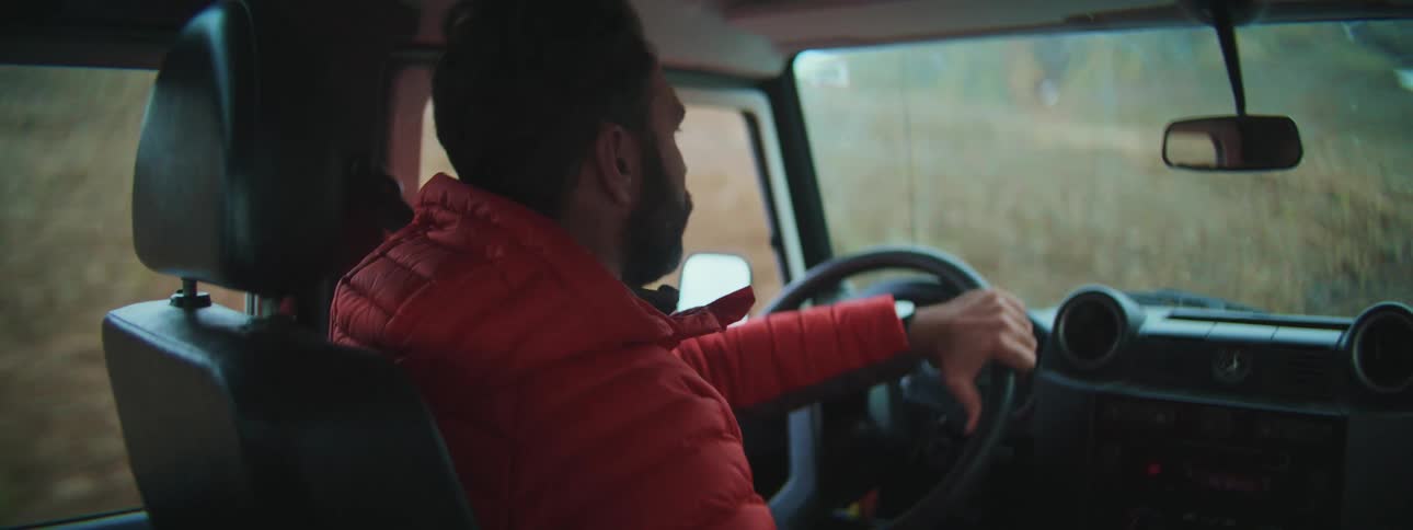 Over-the-shoulder shot of a bearded man in a red jacket driving an off-road vehicle down a rugged country road