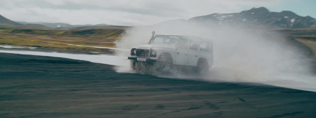 An adventurous white 4x4 drives through a river, splashing water in a rugged Icelandic mountain valley