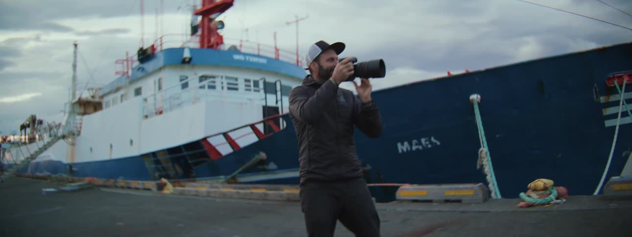 Slow motion shot of a happy photographer walking along a pier, turning around to take pictures with a large boat in the background