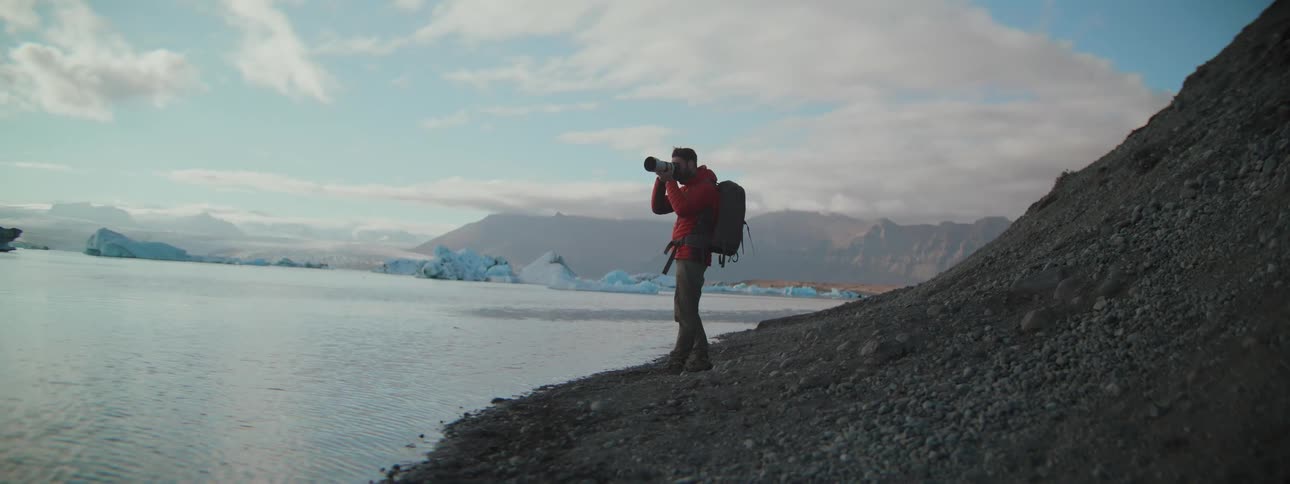 A male photographer in a red jacket stands on a rocky shore and takes pictures of a majestic glacial lagoon